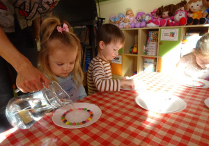 Dziewczynka przygląda się jak pani Ewelina nalewa wodę na talerzyk, chłopiec z prawej stronie układa cukierki na talerzyku a dziewczynka w białej bluzce przygląda się jak rozpuszczają się cukierki.